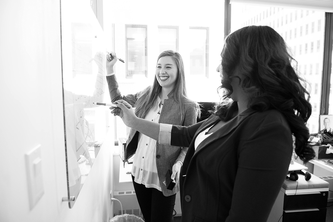 schwarz weiss Fotografie zwei laechelnde junge Frauen vor einem Whiteboard